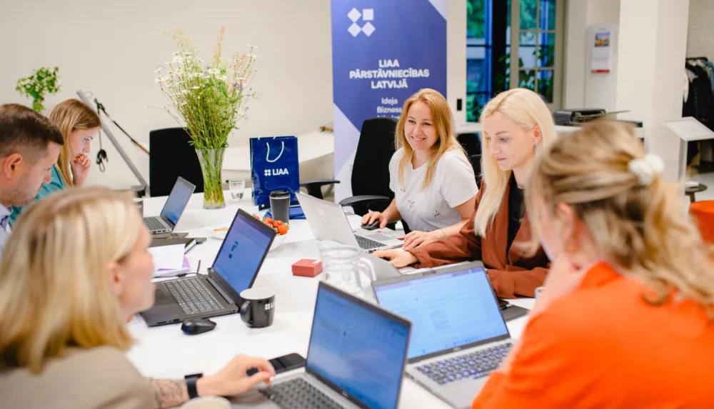 Attēlā var redzēt vairākus cilvēkus, kuri sēž pie datora un piedalās sanāksmē